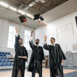 Three male graduates in gowns celebrate with cap toss indoors.